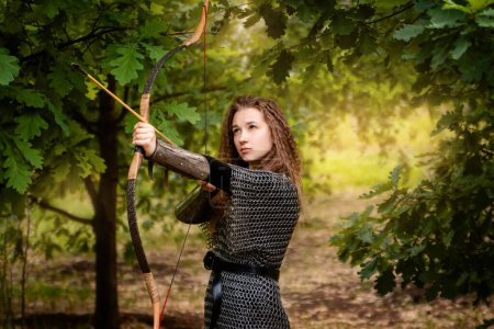 Photo for Medieval warrior woman in chain mail and bracers with a bow in her hands against the backdrop of an evening summer forest. - Royalty Free Image