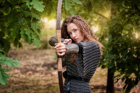 Photo for Medieval warrior woman in chain mail and bracers with a bow in her hands against the backdrop of an evening summer forest. - Royalty Free Image