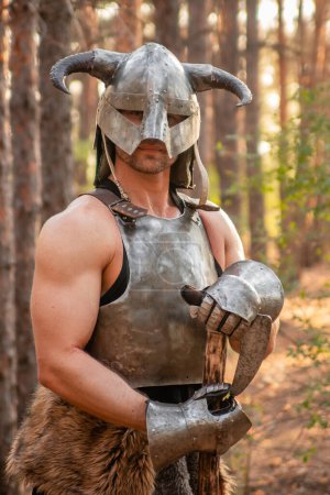 Photo for Portrait of a warrior in a horned helmet, a steel breastplate with a two-handed ax in his hands, posing against the backdrop of the forest. - Royalty Free Image