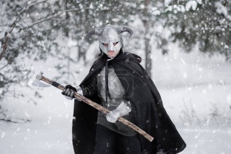 A medieval fantasy warrior in a horned helmet, a steel breastplate and chain mail with a two-handed ax in his hands, walks through snowdrifts in a winter forest.