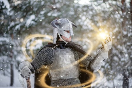 Photo for Portrait of a medieval fantasy warrior in a horned helmet, steel breastplate, chain mail with a two-handed ax in his hand, using a magic spell while standing against the backdrop of a winter forest. - Royalty Free Image