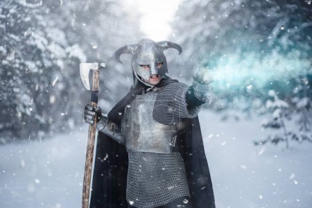 Portrait of a medieval fantasy warrior in a horned helmet, steel breastplate, chain mail with a two-handed ax in his hand, using a magic spell while standing against the backdrop of a winter forest.