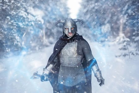 Retrato de un guerrero de fantasía medieval en un casco con cuernos, coraza de acero, cota de malla con un hacha de dos manos en la mano, usando un hechizo mágico mientras está de pie contra el telón de fondo de un bosque de invierno.