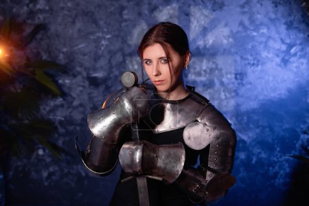 Portrait of a young woman with scars on her face, dressed in black clothes with steel knightly hands and a gorget, holding a bastard sword, posing against an abstract background. Medieval fantasy girl.