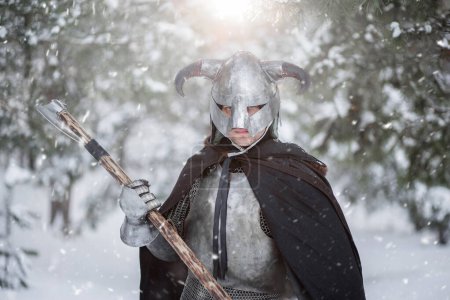 Retrato de un guerrero de fantasía medieval en un casco con cuernos, coraza de acero, cota de malla con un hacha de dos manos en las manos, posando sobre el telón de fondo de un bosque de invierno.