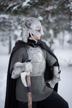 Retrato de un guerrero de fantasía medieval en un casco con cuernos, coraza de acero, cota de malla con un hacha de dos manos en las manos, posando sobre el telón de fondo de un bosque de invierno.