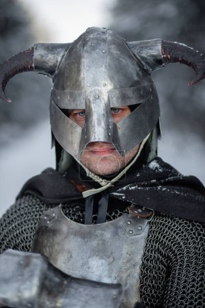 Porträt eines mittelalterlichen Fantasiekriegers mit gehörntem Helm, Stahlpanzer, Kettenhemd mit einer beidhändigen Axt in der Hand, der vor der Kulisse eines Winterwaldes posiert.