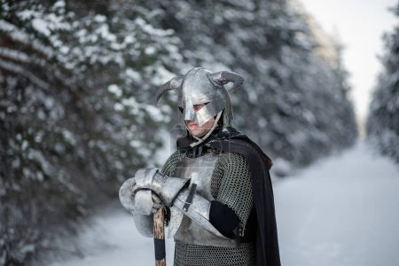 Portrait d'un guerrier fantasme médiéval dans un casque à cornes, cuirasse d'acier, chaîne de courrier avec une hache à deux mains dans ses mains, posant sur le fond d'une forêt d'hiver.
