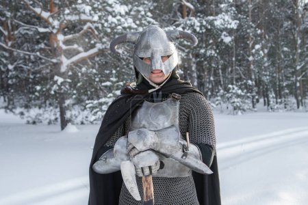 Portrait d'un guerrier fantasme médiéval dans un casque à cornes, cuirasse d'acier, chaîne de courrier avec une hache à deux mains dans ses mains, posant sur le fond d'une forêt d'hiver.