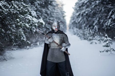 Retrato de un guerrero de fantasía medieval en un casco con cuernos, coraza de acero, cota de malla con un hacha de dos manos en las manos, de pie en una posición de lucha contra el telón de fondo de un bosque de invierno.