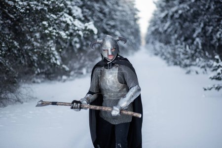 Portrait d'un guerrier fantasme médiéval dans un casque à cornes, cuirasse en acier, chaîne de courrier avec une hache à deux mains dans ses mains, debout dans une position de combat dans le contexte d'une forêt d'hiver.