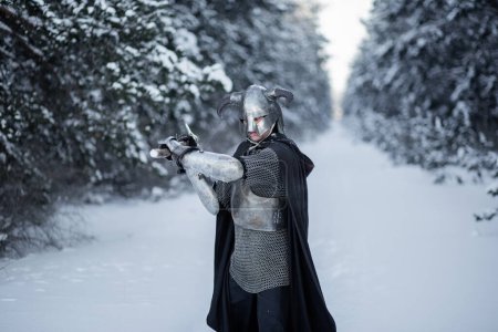 Retrato de un guerrero de fantasía medieval en un casco con cuernos, coraza de acero, cota de malla con un hacha de dos manos en las manos, de pie en una posición de lucha contra el telón de fondo de un bosque de invierno.