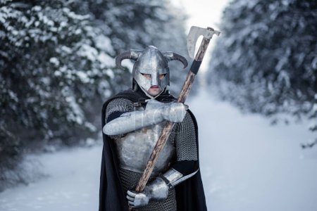 Porträt eines mittelalterlichen Fantasiekriegers mit gehörntem Helm, Stahlpanzer, Kettenpanzer mit einer beidhändigen Axt in der Hand, der vor der Kulisse eines Winterwaldes in Kampfstellung steht.