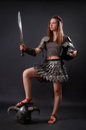Full-length portrait of a medieval woman warrior in female armor with a sword in her hand standing in a heroic pose with her foot on a horned helmet isolated on a dark background.