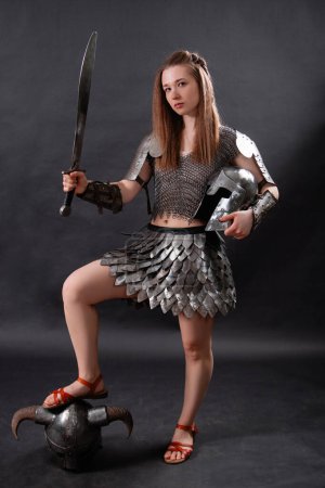 Ganzkörperporträt einer mittelalterlichen Kriegerin in weiblicher Rüstung mit einem Schwert in der Hand, die in heldenhafter Pose mit dem Fuß auf einem gehörnten Helm vor dunklem Hintergrund steht..