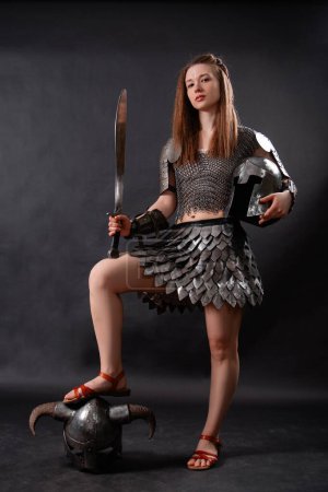 Ganzkörperporträt einer mittelalterlichen Kriegerin in weiblicher Rüstung mit einem Schwert in der Hand, die in heldenhafter Pose mit dem Fuß auf einem gehörnten Helm vor dunklem Hintergrund steht..