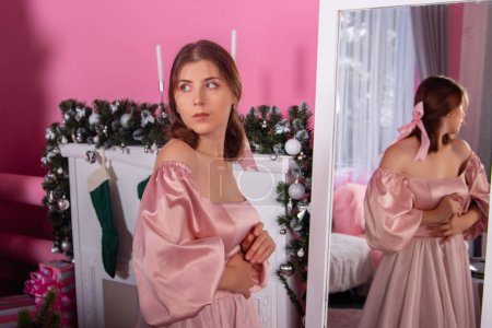 Portrait of a young woman in a pink dress standing near a mirror against the backdrop of Christmas decorations.