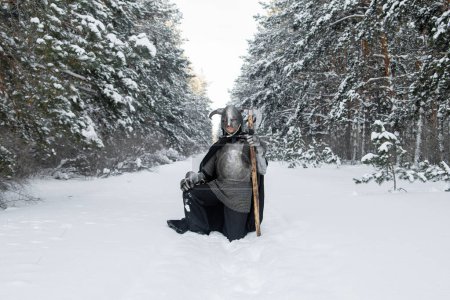 Full-length portrait of a medieval fantasy warrior in a horned helmet, steel breastplate, chain mail with a two-handed ax in his hands, posing while sitting against the backdrop of a winter forest.