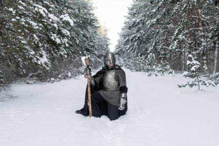 Ganztägiges Porträt eines mittelalterlichen Fantasiekriegers mit gehörntem Helm, stählernen Brustpanzer, Kettenhemd mit einer beidhändigen Axt in den Händen, der im Sitzen vor der Kulisse eines Winterwaldes posiert.