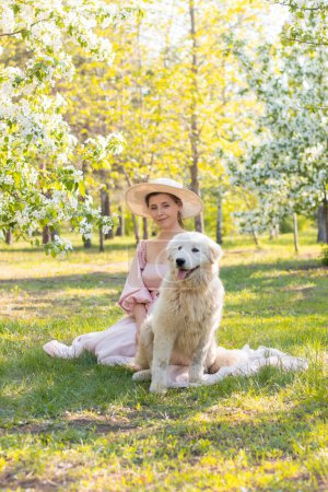 Portrait of a young woman dressed in a pink dress with a train and a hat, sitting next to a large white fluffy dog against the backdrop of a spring park and blooming flowers.