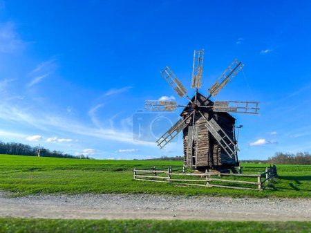 Rustikale hölzerne Windmühle in idyllischer Landschaft. Ein faszinierendes Bild, das eine traditionelle hölzerne Windmühle mit ihren verwitterten Segeln vor einem strahlend blauen Himmel zeigt. Die Szene ist auf einem saftig grünen Feld komponiert.