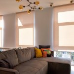 Motorized roller blinds. A sofa with colorful pillows in the room near windows with roller blinds. Automatic roller shades on large window to the floor in the interior. Sunny day. Selective fokus.