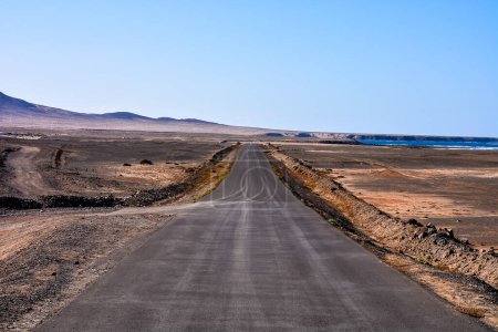 A long road stretches across a desert landscape. The sky is clear and blue, and the road is empty