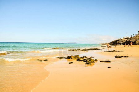 Ein Strand mit einem klaren blauen Meer und ein paar Menschen, die auf dem Sand spazieren gehen. Der Strand ist felsig und das Wasser ist ruhig