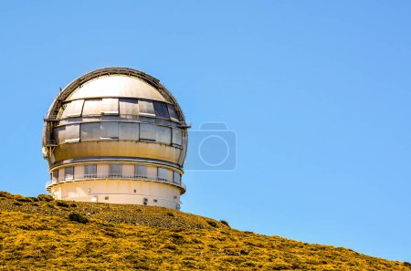 A large telescope is on a hill with a clear blue sky. The telescope is surrounded by a grassy hillside