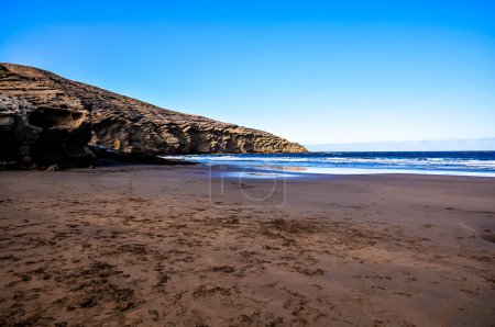 Une plage avec une falaise rocheuse en arrière-plan. Le ciel est clair et bleu