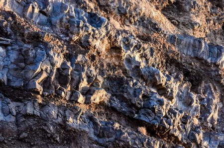 Das Bild zeigt eine felsige Klippe mit einem blauen und grauen Farbschema. Die Felsen sind zerklüftet und rau, was den Eindruck einer rauen und zerklüfteten Umgebung vermittelt. Das Sonnenlicht scheint auf die Felsen