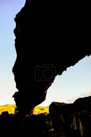 A large rock formation with a dark shadow cast on it. The sky is blue and the sun is setting