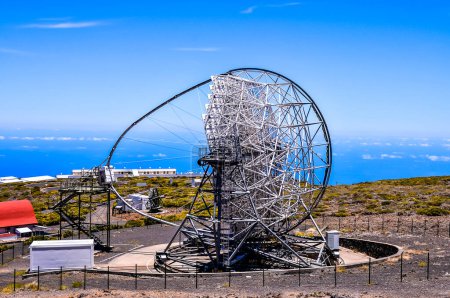 Ein großes Teleskop steht auf einem Hügel mit Blick auf den Ozean. Das Teleskop ist von einem Zaun umgeben und von einem roten Gebäude umgeben. Der Himmel ist klar und blau, und der Ozean ist in der Ferne sichtbar