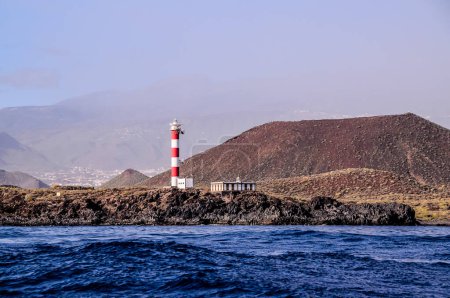 A lighthouse is on a rocky island in the ocean. The lighthouse is red and white. The ocean is calm and the sky is clear, real image