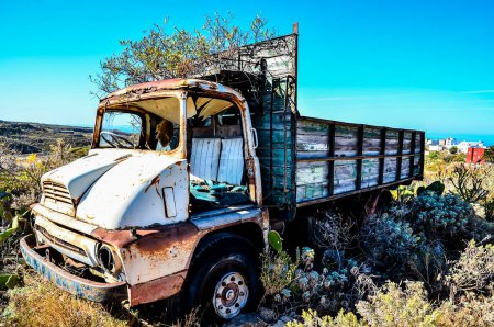 An old truck is sitting in a field with a blue sky in the background. The truck is rusted and has a broken windshield, real image