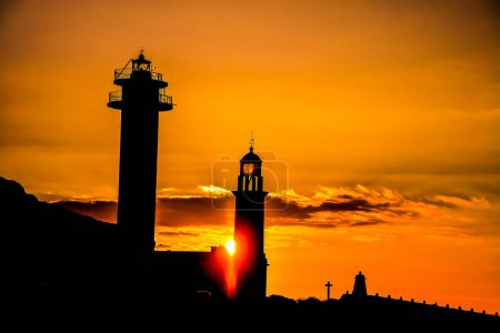 A lighthouse and a small building are silhouetted against a sunset. The sun is setting and the sky is orange