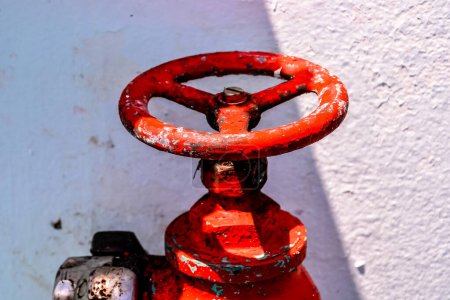 Ein roter Feuerhydrant mit einem verrosteten und schmutzig aussehenden Griff. Der Hydrant steht an einer weißen Wand
