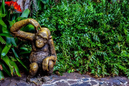 A bronze statue of a monkey is sitting on a stone path next to some green plants. The statue is looking to the left, as if it is pondering something. The scene has a calm and peaceful atmosphere