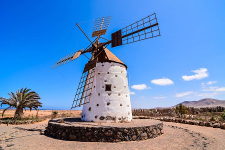 A windmill is standing in a field with a clear blue sky above it. The windmill is old and has a rustic appearance. The scene is peaceful and serene, with the windmill as the focal point