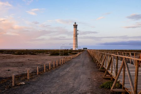 Ein Leuchtturm steht auf einem Steg vor einem Strand. Der Himmel ist bewölkt und die Sonne geht unter