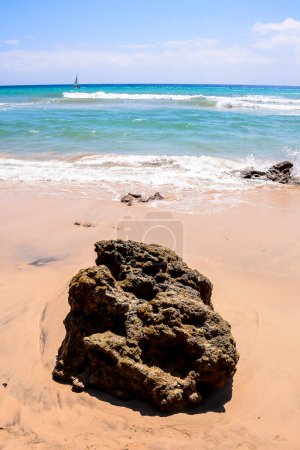 Ein großer Felsen liegt am Strand, im Hintergrund das Meer. Die Szene ist friedlich und heiter, die Wellen plätschern sanft am Ufer entlang.