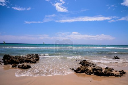 Une belle plage avec un ciel bleu clair et quelques rochers dans l'eau. L'eau est calme et le ciel est lumineux et ensoleillé