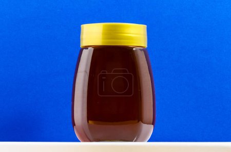 Un frasco de miel está sentado en una superficie azul. El frasco está hecho de vidrio y tiene una tapa de oro. La miel dentro del frasco es marrón y él es grueso