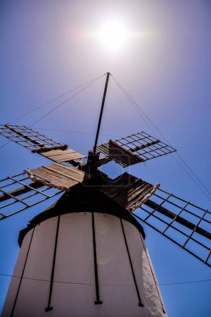 A windmill is standing tall in the sky with the sun shining on it. Concept of strength and stability, as the windmill stands tall and proud against the blue sky. The sun's rays illuminate the windmill