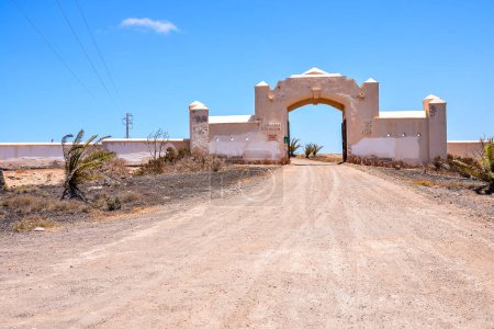 A dirt road leads to a large white building with a gate 