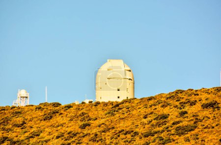 Un gran telescopio blanco está en una colina con un cielo azul claro. El telescopio es el foco principal de la imagen, y la colina y el cielo proporcionan un telón de fondo sereno y pacífico