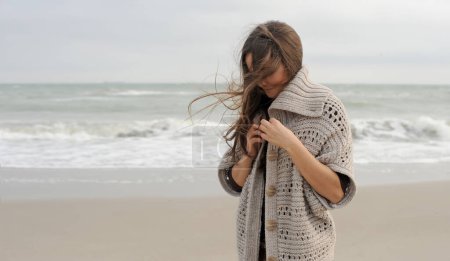 Foto de Young brunette woman portrait dressed knitted sweater, hiking near the ocean, windy weather - Imagen libre de derechos