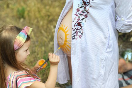 Foto de Niña pintando sol en el vientre de mamá embarazada, al aire libre en el parque - Imagen libre de derechos