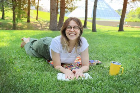 Téléchargez les photos : Heureuse étudiante se reposer dans un parc sur une herbe avec un livre, regardant la caméra - en image libre de droit