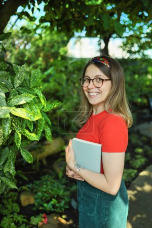 Téléchargez les photos : Jeune étudiante étudiant des plantes dans un jardin botanique, regardant la caméra et souriant - en image libre de droit